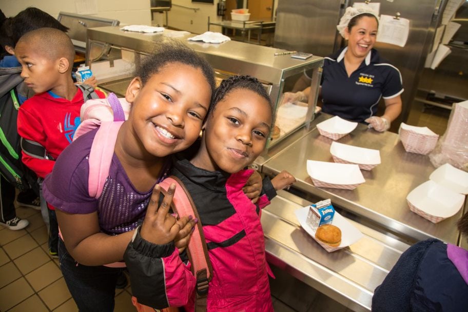 Two little elementary school girls in the cafeteria serving line with big smiles while a school nutrition professional smiles from behind the line